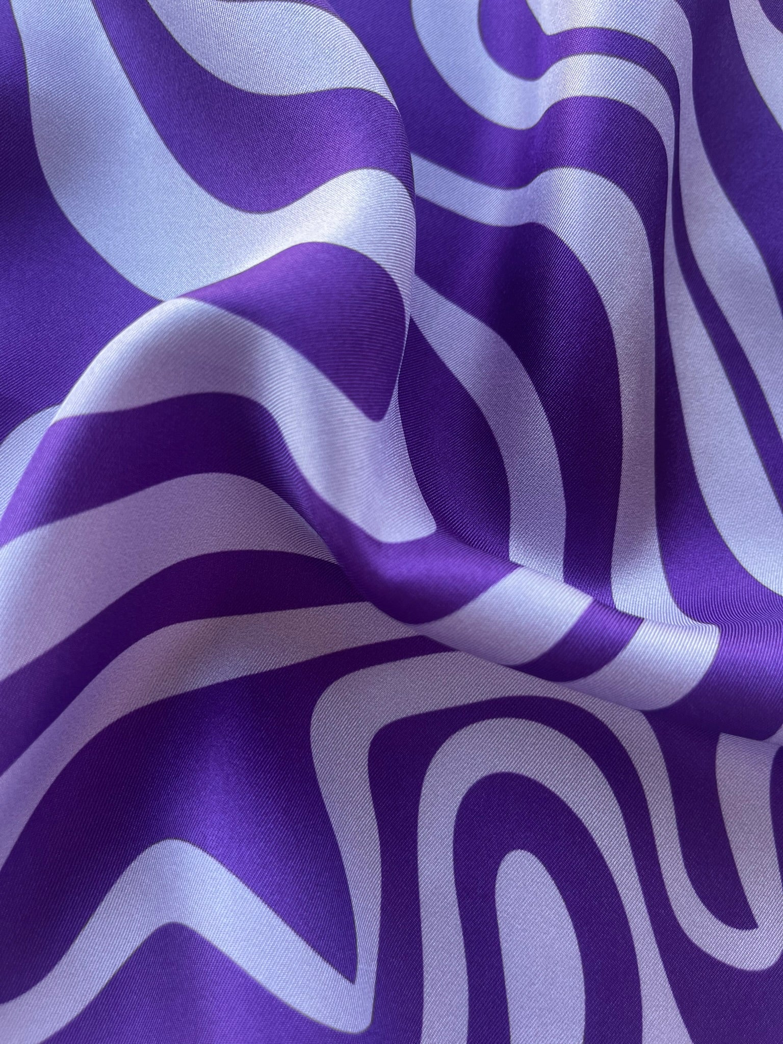 Close-up detail of the luxurious silk fabric texture of the apashka Acid Wave silk scarf, highlighting the high-shine material and the bold purple and light lavender psychedelic wavy pattern.