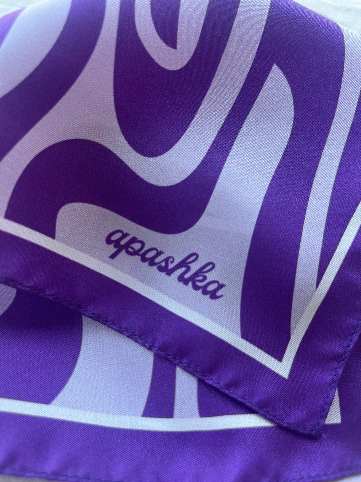 Close-up of the folded corner of the apashka Acid Wave silk scarf, showcasing the dark purple cursive brand logo printed over a lavender and violet abstract swirl pattern.