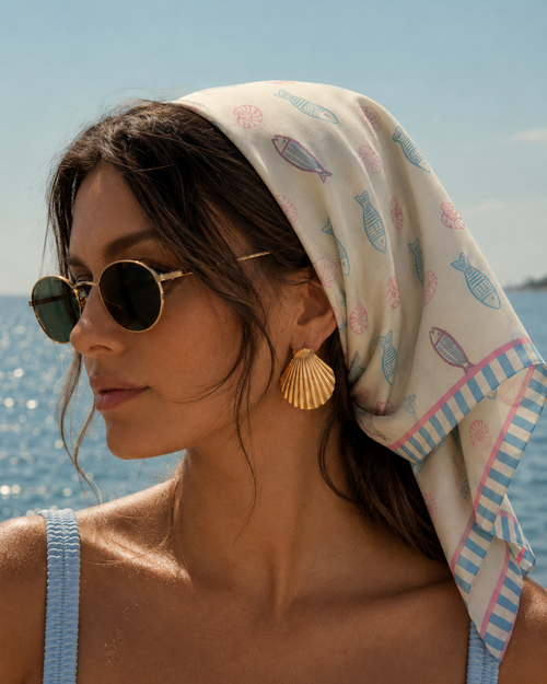 A model at sea wearing the apashka Marina silk bandana as a headscarf, styled with sunglasses and gold shell earrings.