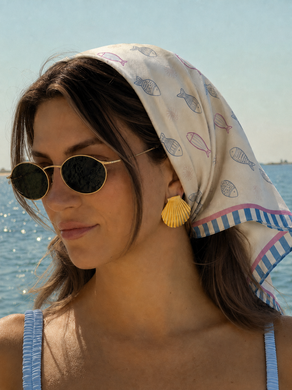 A model at sea wearing the apashka Marina silk bandana as a headscarf, styled with sunglasses and gold shell earrings.
