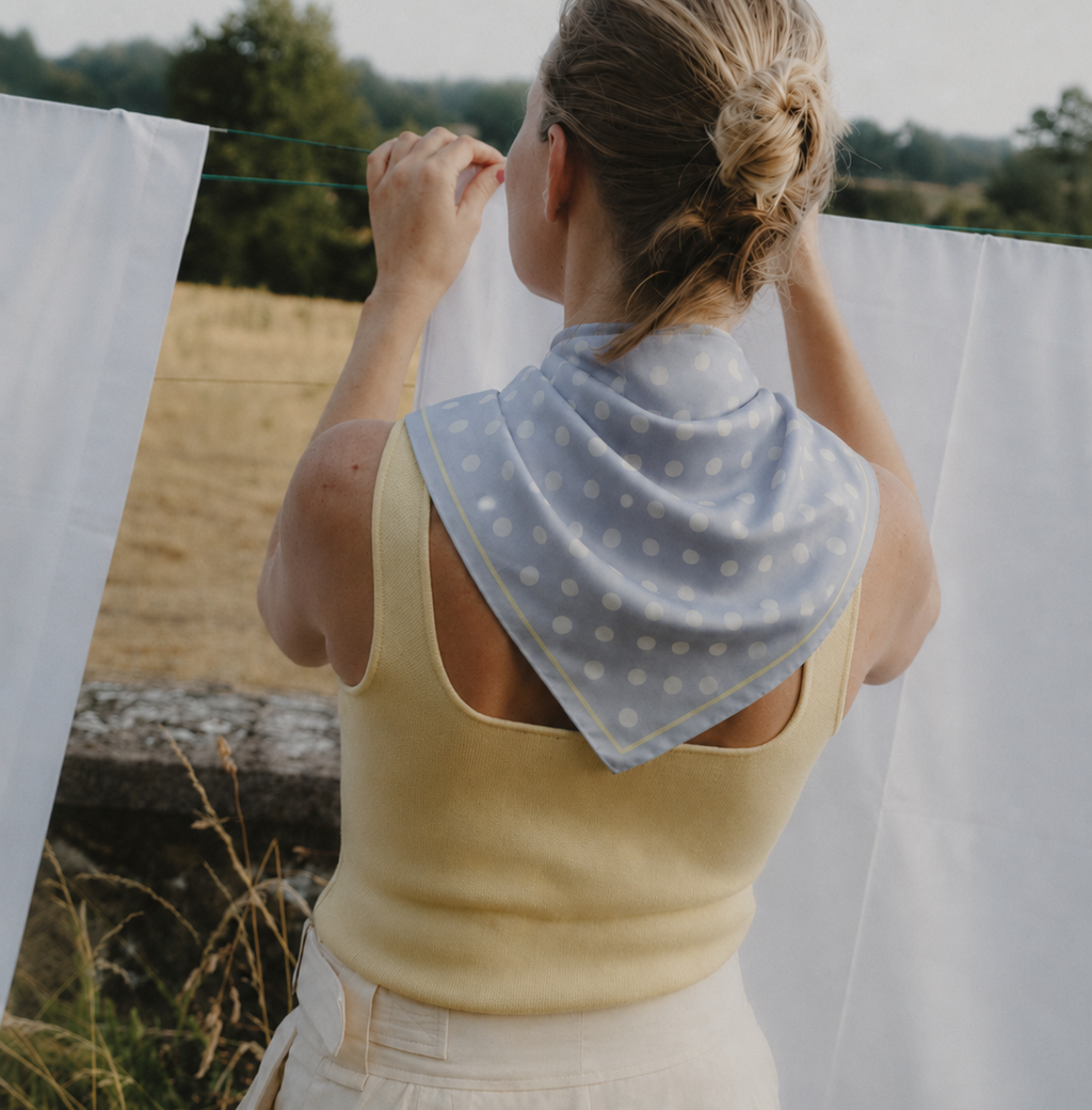 Rear view of a model styling the apashka Bubble-Pop Blu polka dot silk scarf tied around her neck, paired with a yellow tank top in a rustic outdoor setting.