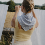 Rear view of a model styling the apashka Bubble-Pop Blu polka dot silk scarf tied around her neck, paired with a yellow tank top in a rustic outdoor setting.