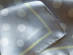 Close-up of the corner of the apashka Bubble-Pop Blu silk scarf, showing the white cursive brand logo on light blue silk fabric with white polka dots and a pale yellow inner border line.