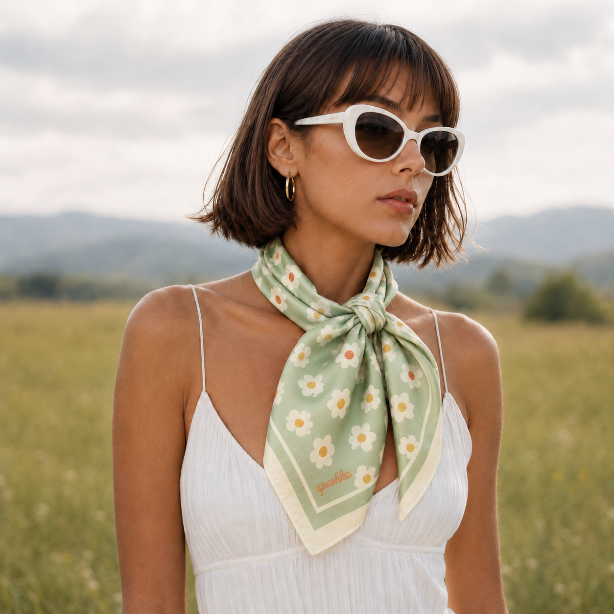 A model wearing the apashka Daisy Daze 100% silk bandana tied as a front-knot neckerchief, styled with a white summer dress and retro sunglasses to highlight the sage green daisy floral print.