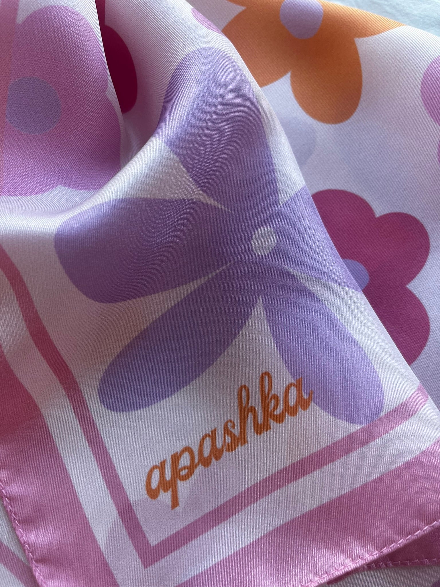 Close up of the signature apashka script logo on the baby pink border corner of the retro floral silk bandana.