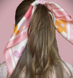 Rear view of a model styling the apashka Fiori Fuori floral silk scarf tied as a retro hair accessory around a ponytail, showcasing its pink, orange, and lavender flower pattern against a pink background.