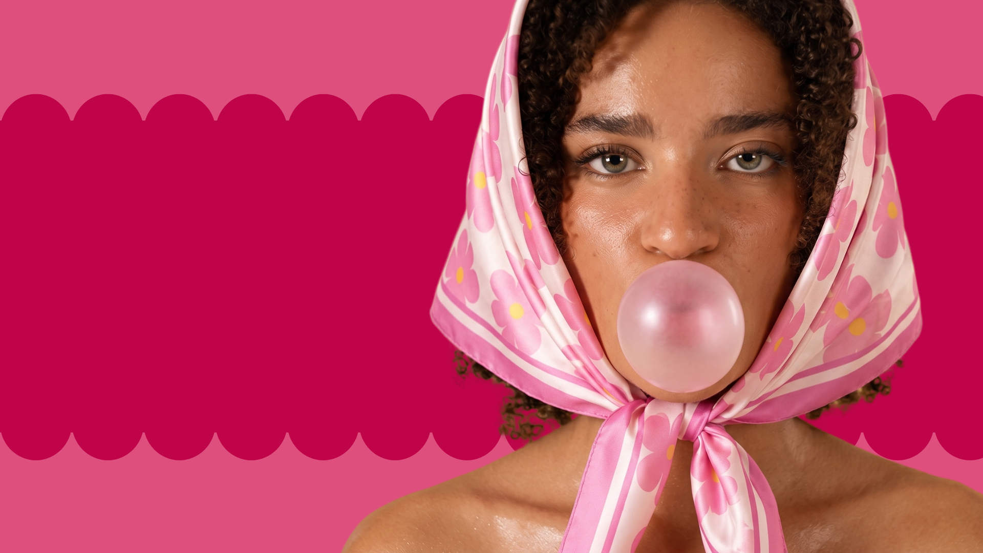 Woman blowing a pink bubble gum bubble with a pink floral headscarf against a pink background