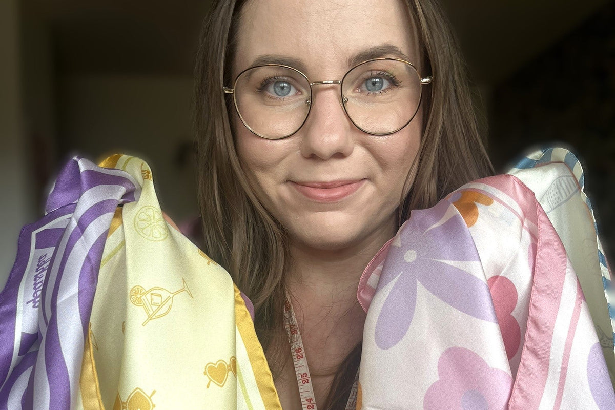 A woman with glasses holding up two patterned silk scarves. The scarf on the left is pale yellow with gold cocktail glass motifs and purple striped borders.