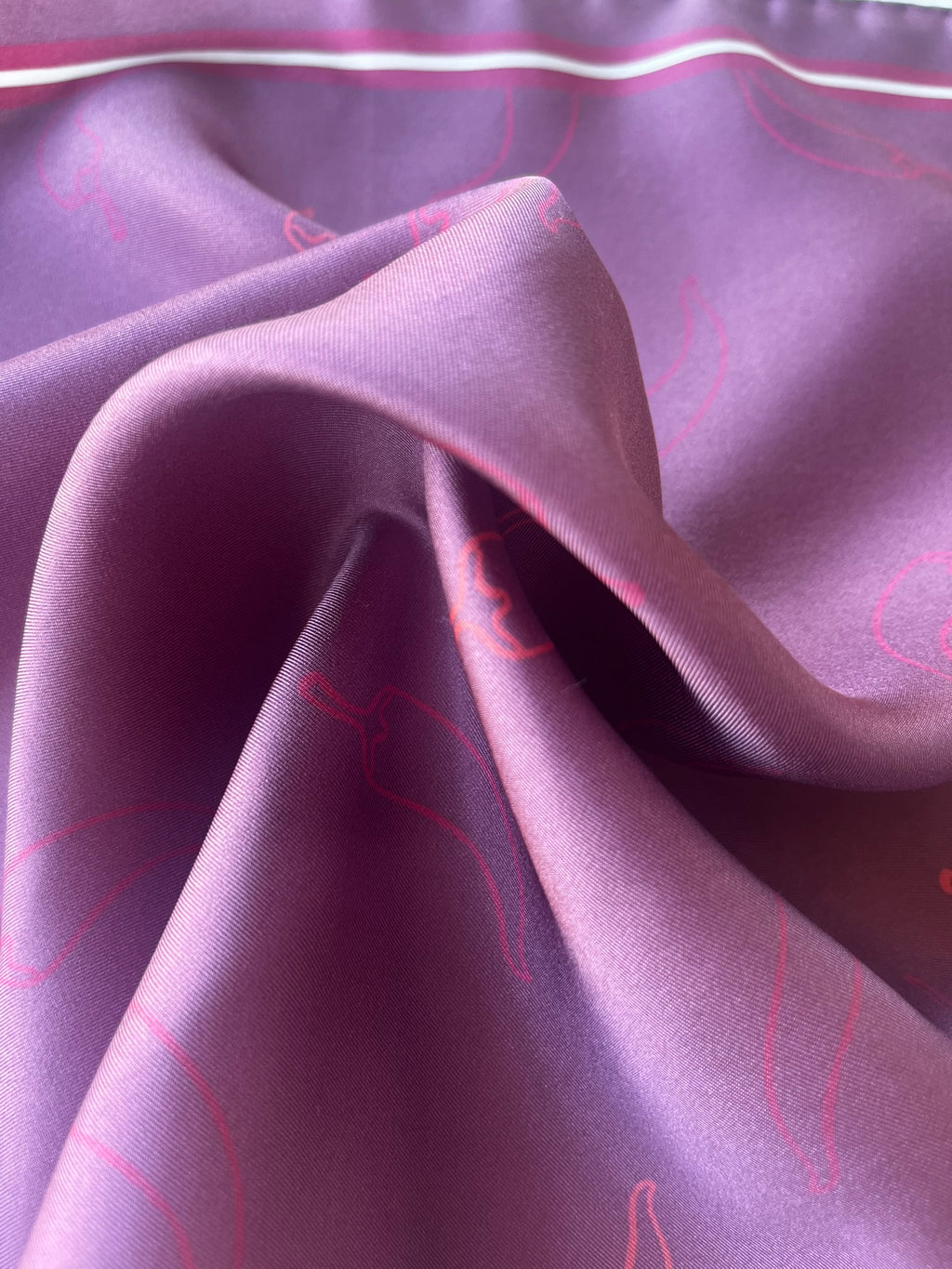 A close-up of draped purple fabric with a subtle pink chili pepper pattern and a thin white stripe along the top edge.