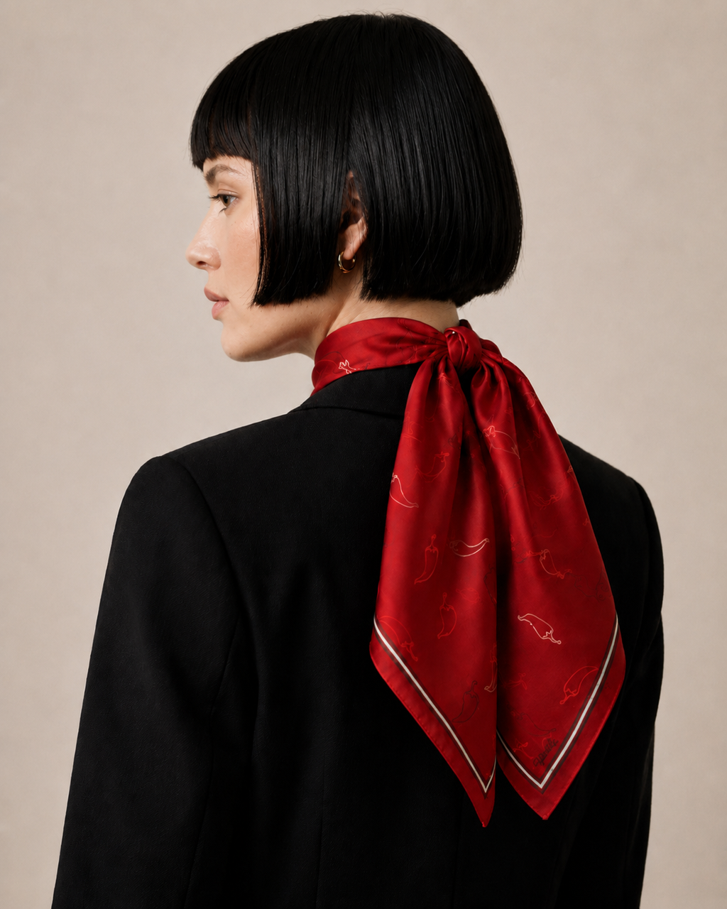 A rear view of a model wearing the apashka Red Hot Chili 100% silk bandana tied securely at the back of the neck, styled with a sharp black blazer and showcasing the crimson chili pepper print pattern.