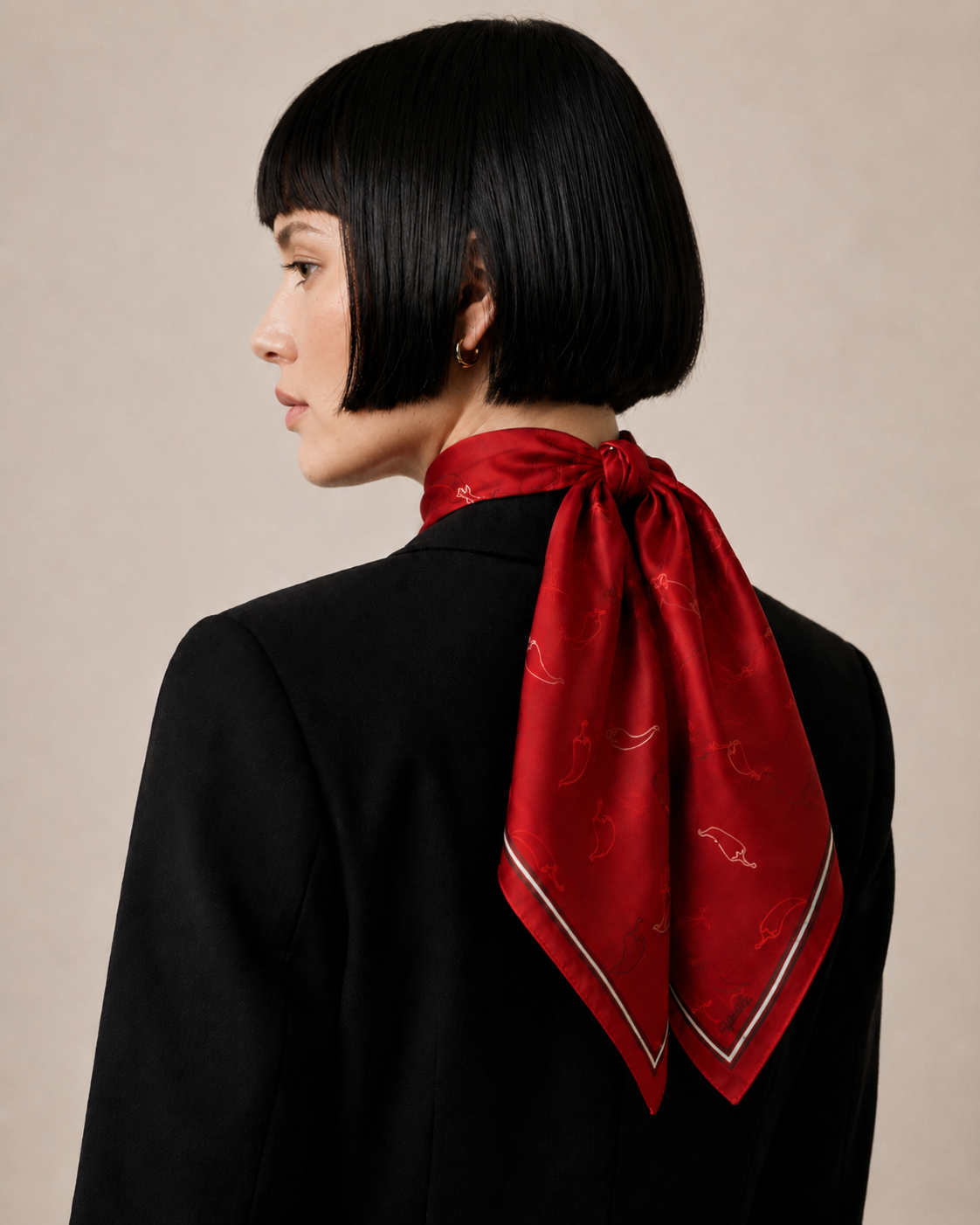 A rear view of a model wearing the apashka Red Hot Chili 100% silk bandana tied securely at the back of the neck, styled with a sharp black blazer and showcasing the crimson chili pepper print pattern.