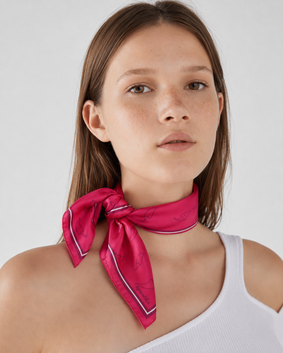 Model wearing the apashka Red Hot Chili Apashka crimson silk bandana, tied as a front-knot neckerchief to showcase the chili pattern and border, paired with a white tank top.