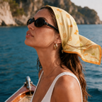 A model on a wooden boat at sea wearing the apashka Sole e Spritz yellow silk bandana as a retro headscarf, styled with black sunglasses and a white top.