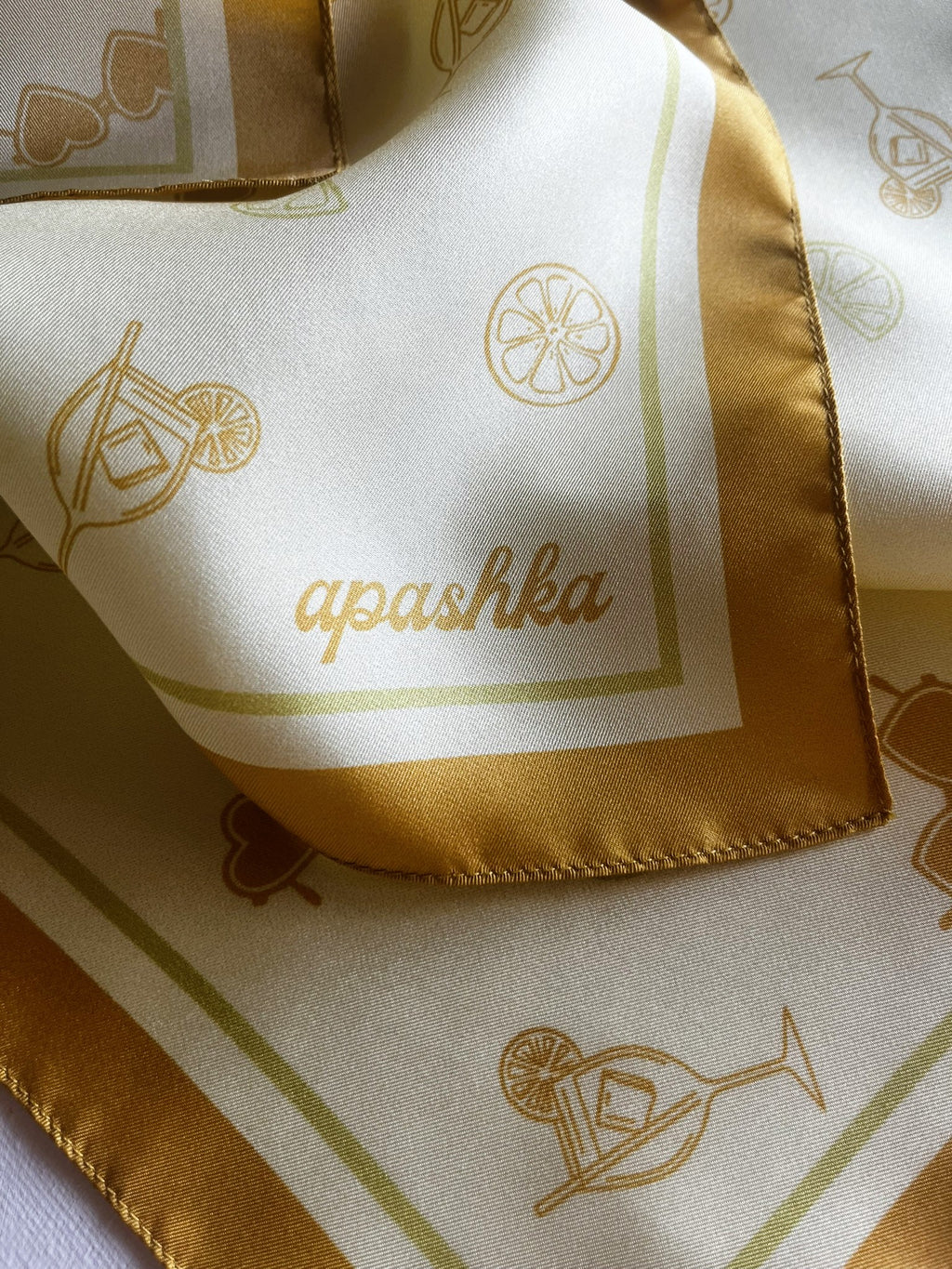 A close-up shot of a folded, lustrous cream silk scarf highlighting the brand name "apashka" printed in a cursive font. The fabric features delicate line drawings of cocktail glasses, citrus wheels, and heart-shaped sunglasses, bordered by a thick golden-orange hem.