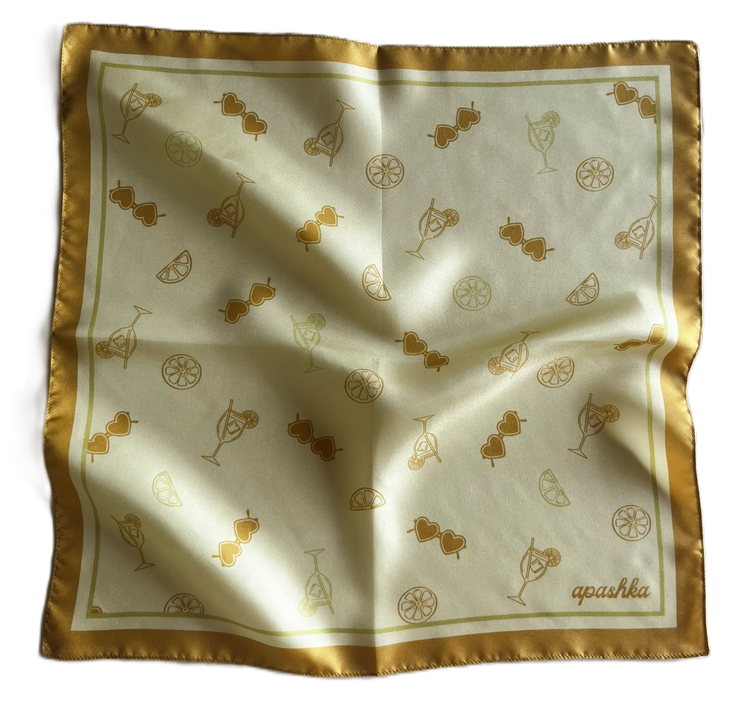 A top-down view of a square silk scarf slightly rippled and draped. The scarf has a warm cream-colored center scattered with an all-over gold orange art pattern of cocktails, lemon slices, and heart sunglasses, framed by a thick golden-yellow border.