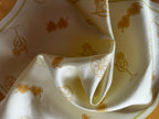 A detailed close-up of the smooth, shiny texture of a cream-colored silk fabric twisted into an elegant swirl. The fabric is printed with gold outline illustrations of summer-themed motifs including heart-shaped sunglasses, cocktail glasses, and sliced citrus fruits.