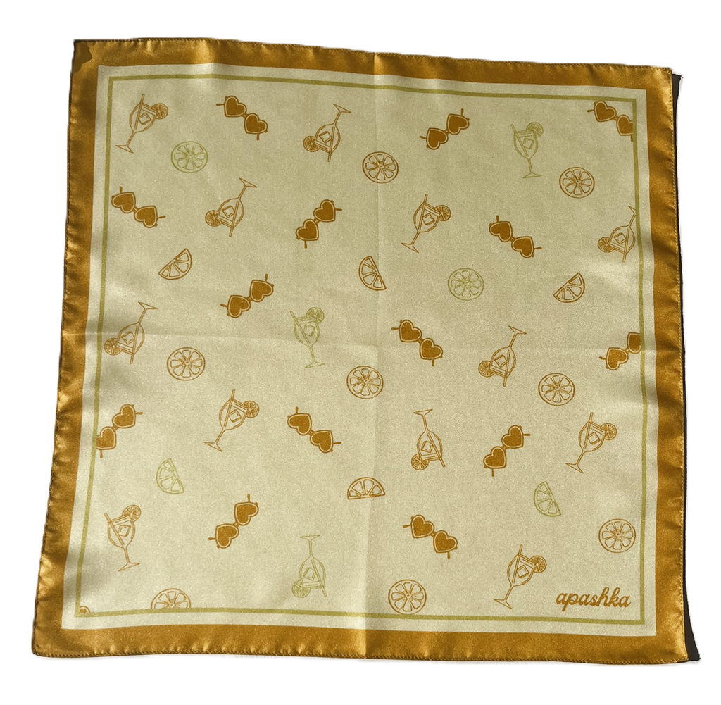 A flat, full-view layout of silk scarf. The scarf displays a repeated pattern of gold orange line-drawn cocktails, citrus wheels, and heart-shaped sunglasses, finished with a solid golden-orange border and the brand name "apashka" in the lower right corner.