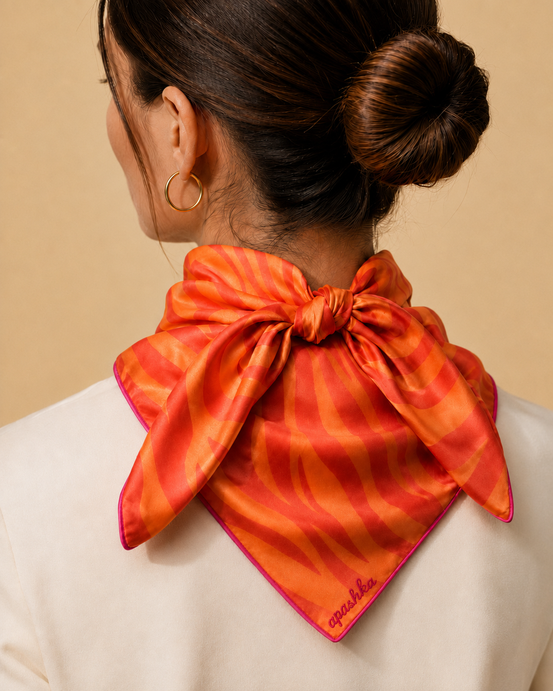 A model viewed from behind with her hair in a sleek bun, styling the apashka Sunset Safari orange and tangerine zebra print silk bandana tied as a neckerchief over a cream blazer, prominently displaying the signature contrast hot pink hand-rolled hem.
